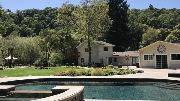 A two-story building with a clock on the side, surrounded by lush greenery and a swimming pool.