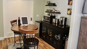A compact kitchen with a wooden dining table, chairs, a refrigerator, microwave, and a coffee maker. There is a chalkboard sign on the wall and a shelf with kitchen items.