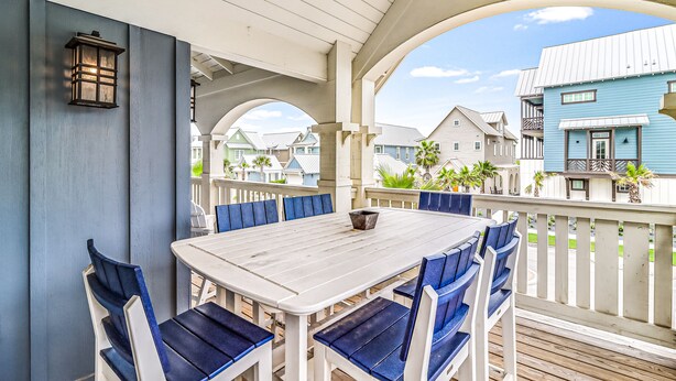 Een overdekte eethoek buiten met een witte tafel en blauwe stoelen, omringd door een witte hek en met uitzicht op een woonwijk.