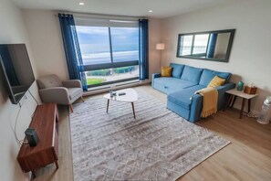 Ein modernes Wohnzimmer mit einer Couch, einem Couchtisch, einem Fernseher und einem großen Fenster mit Blick ins Freie.