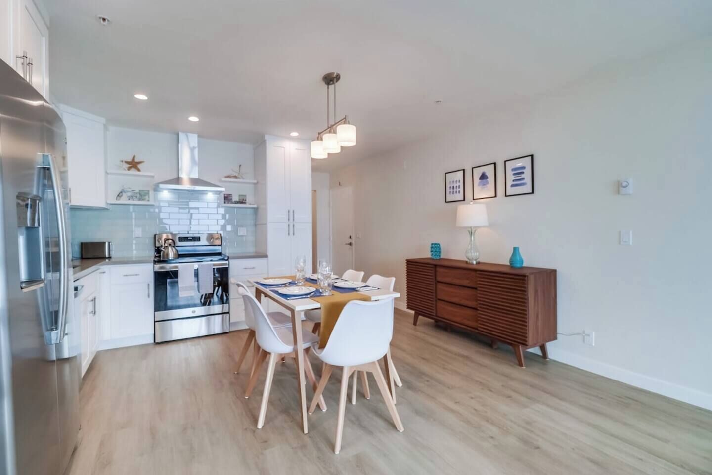 A modern kitchen with stainless steel appliances, a dining area with a wooden table and white chairs, and a wooden cabinet with decorative items.