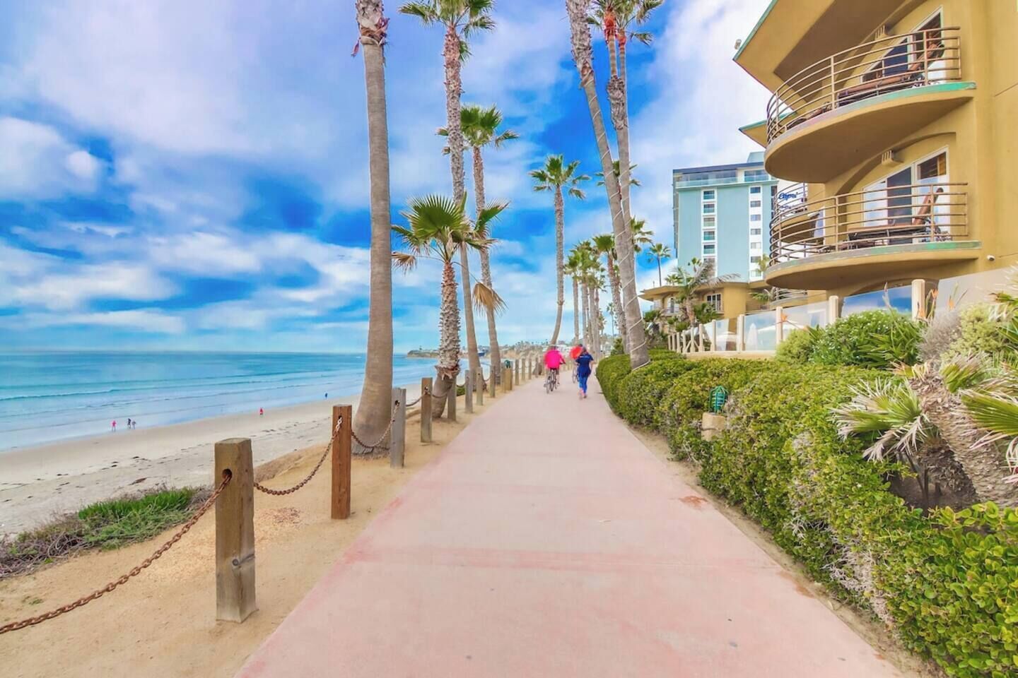 A beachfront walkway lined with palm trees and a building with balconies overlooking the ocean.
