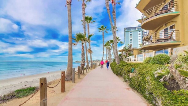 A beachfront walkway lined with palm trees and a building with balconies overlooking the ocean. - Cozy Oceanfront view, beach access, pool, parking (San Diego)