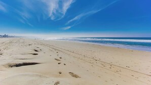 A beach with footprints leading to the ocean, a clear sky, and a distant city skyline. - Cozy Oceanfront view, beach access, pool, parking (San Diego)