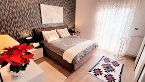 A bedroom with a bed, bedside lamps, a nightstand, a potted plant, a patterned rug, and a window with curtains. - Villa Etna, with swimming pool and garden.  (Blandano)