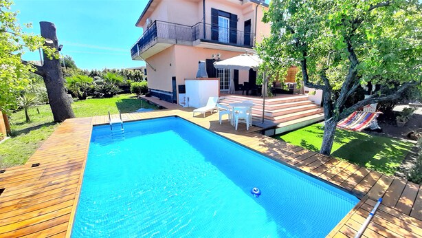 A rectangular swimming pool with a wooden deck, surrounded by a well-maintained lawn and a two-story house with a balcony. - Villa Etna, with swimming pool and garden.  (Blandano)