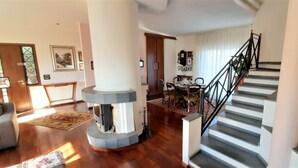 A spacious living room with a wooden floor, a large column, a staircase with black railings, and a dining area with chairs and a table. - Villa Etna, with swimming pool and garden.  (Blandano)