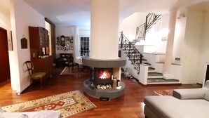 A spacious living room with a fireplace, wooden flooring, and a staircase leading to an upper level. - Villa Etna, with swimming pool and garden.  (Blandano)