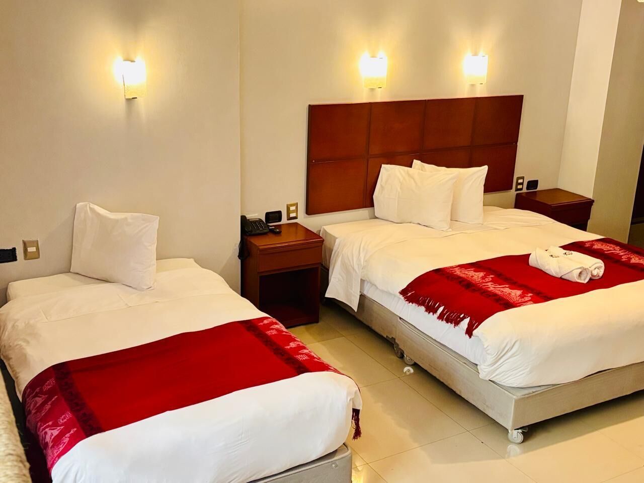 Habitación de hotel con dos camas individuales, cada una con cabecero de madera, ropa de cama blanca y una manta roja. Se observan mesitas de noche con lámparas y un pequeño aparador con un teléfono.