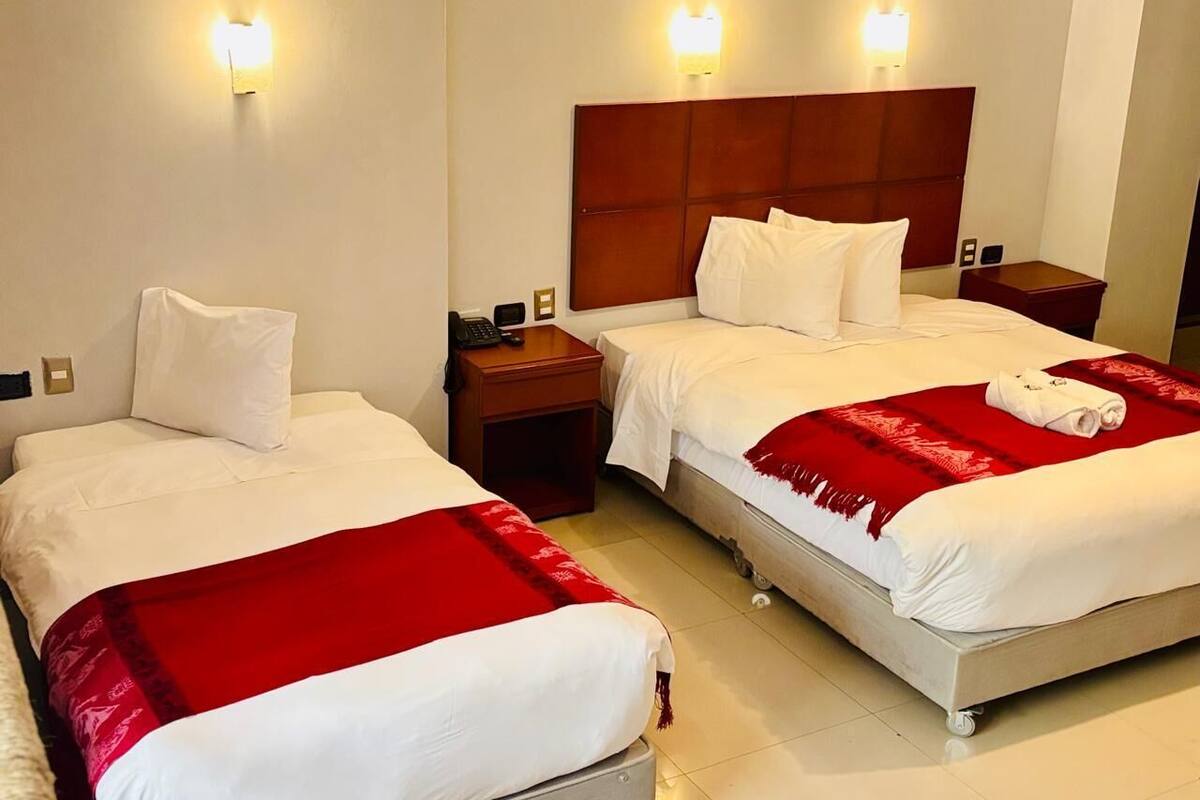 A hotel room with two single beds, each with a wooden headboard, white bedding, and a red throw blanket. There are bedside tables with lamps and a small nightstand with a phone.