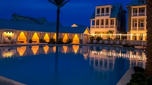 Una piscina con el reflejo de edificios y luces, rodeada de sillas de descanso y palmeras.
