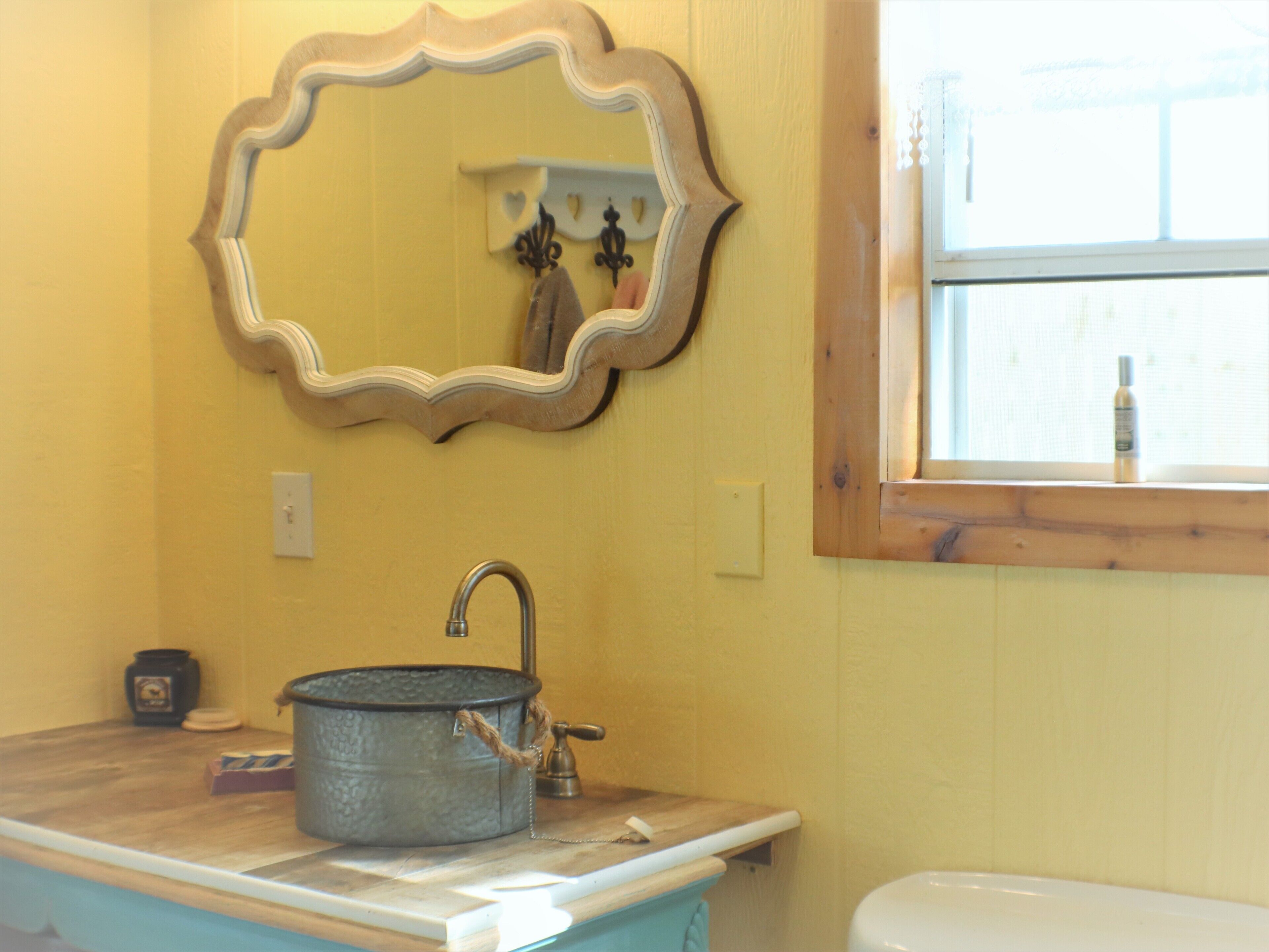 A bathroom with a yellow wall, a wooden-framed mirror, a sink with a metal basin, and a window.