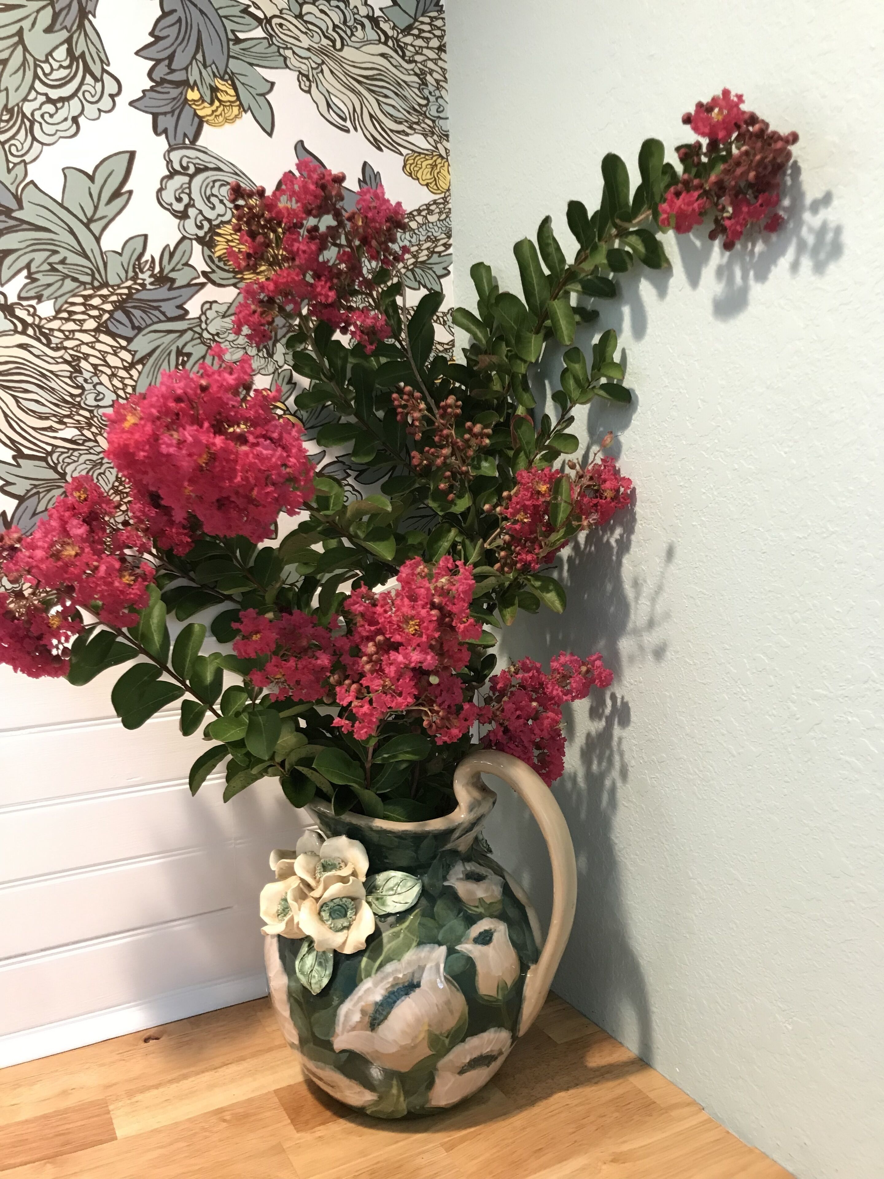A vase with pink flowers and a floral design, placed on a wooden floor against a wall with a patterned design.