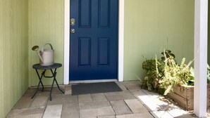 Une porte bleue, avec un paillasson devant et une petite table à côté, sur laquelle se trouve un arrosoir.