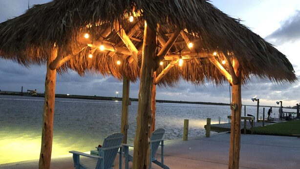 A wooden gazebo with a thatched roof and string lights, overlooking a body of water.