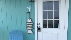 A teal-painted exterior with a white door, a blue Adirondack chair with a star-patterned pillow, a welcome mat, and a sign reading "Welcome to the Beach."