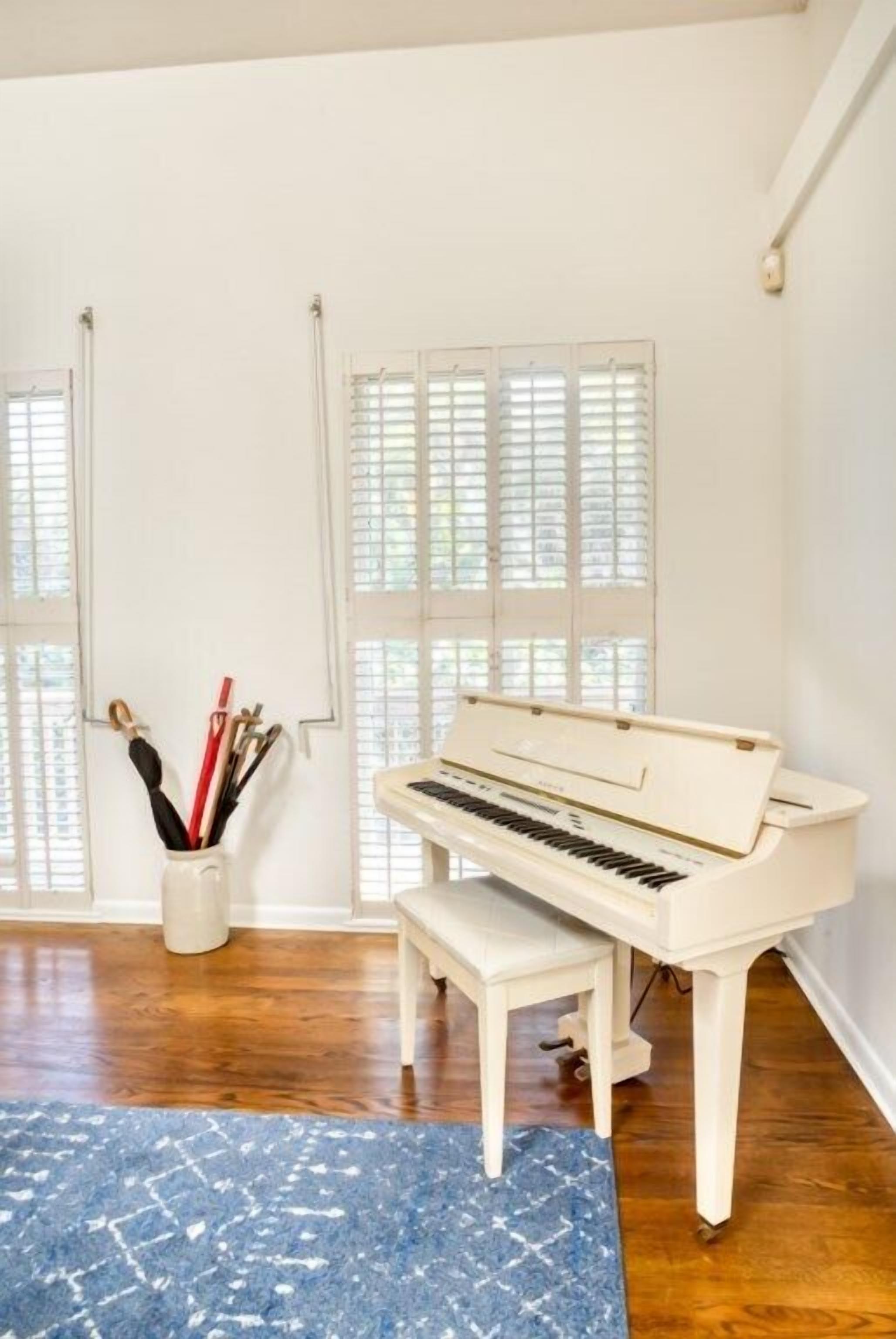 Un piano en una habitació amb terres de fusta i parets blanques, una catifa blava i una finestra amb persianes.