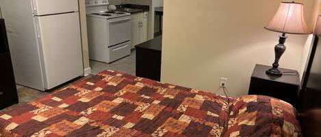 A bedroom with a bed, a nightstand with a lamp, a kitchenette with a refrigerator, stove, and oven.