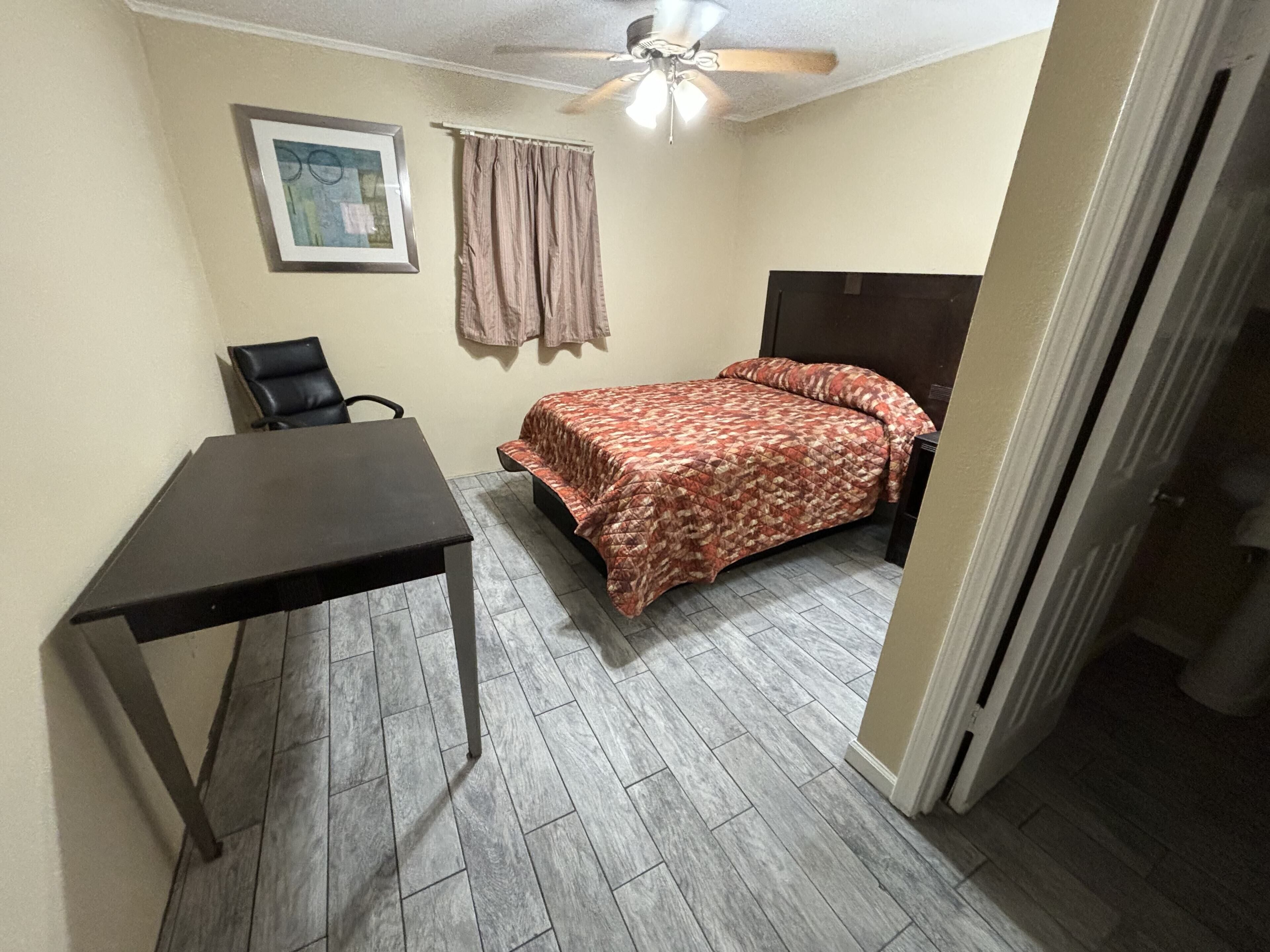 A small, well-lit room with a bed, a desk, and a chair.