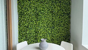 Ein Zimmer mit einem runden weißen Tisch, einer grauen Vase und weißen Stühlen. Eine grüne, blätterreiche Wand befindet sich dahinter.
