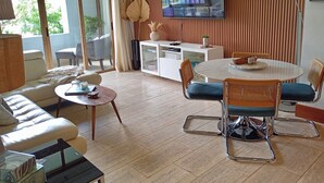 Ein modernes Wohnzimmer mit einer Couch, einem runden Tisch und Stühlen. Ein Fernseher ist an der Wand montiert, es gibt eine Lampe und eine Kommode mit Dekoration.