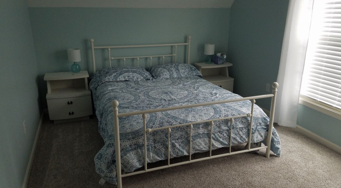 Quarto com estrutura de cama em metal, mesa de cabeceira com abajur e mesinha de apoio com luminária.