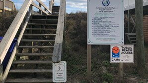 Een houten trap die leidt naar een strandgedeelte, met een bord waarop de regels voor toegang tot het strand staan en een logo van het Coastal Management Program.