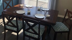 Een eetruimte met een houten tafel gedekt voor vier personen, stoelen met gestreepte kussens, een kroonluchter en uitzicht op een balkon met de tekst 'RELAX NOW YOU'RE AT THE BEACH'.