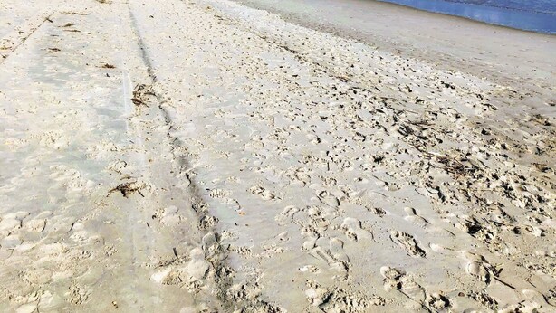 Een strand met voetafdrukken, een houten constructie en uitzicht op de oceaan onder een bewolkte lucht.