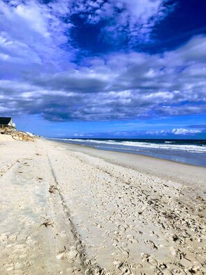 Ein Strand mit Fußspuren, einer Holzkonstruktion und Blick auf den Ozean unter einem teils bewölkten Himmel.