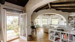 Una habitación amplia con un gran ventanal abierto que da a un balcón, una zona de estar acogedora con un sofá y una mesa, y una estantería llena de libros.