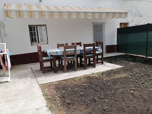 Un espace extérieur abrité, aménagé pour les repas, avec une table et des chaises, un petit jardin et un bâtiment doté d’une fenêtre et d’une porte.