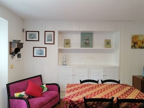 Une pièce avec un canapé violet, une table à manger recouverte d’une nappe à motifs et des lambris blancs sur les murs.