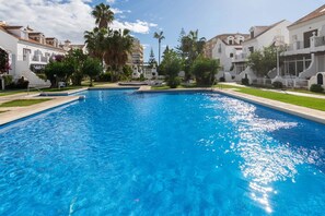 A swimming pool surrounded by white buildings with balconies, palm trees, and a clear sky.