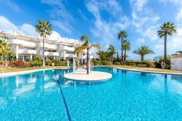A swimming pool with a circular island, palm trees, and a building with balconies in the background.