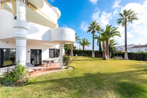 A modern two-story house with a balcony, a patio with chairs, and a well-maintained lawn with palm trees.