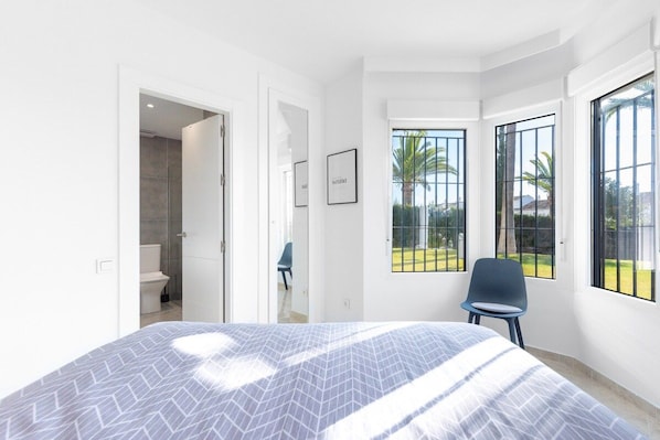 A bedroom with a bed, a chair, and a view of the outdoors through large windows. - Antares I | 2BR Garden & Pool View Apartment in Calahonda Near Marbella (Mijas Costa)