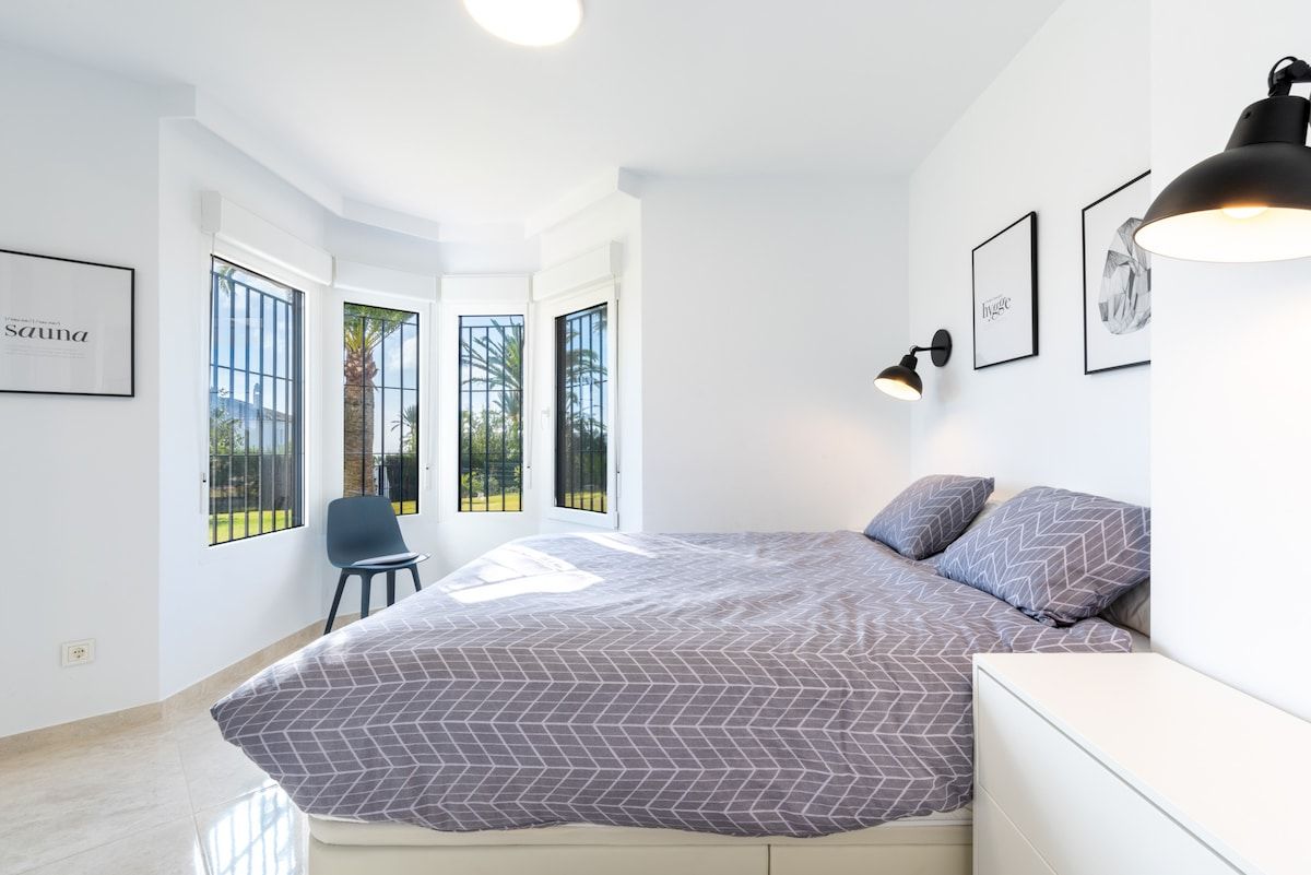 A modern bedroom with a large bed, a chair, and a desk. There are windows with a view of greenery outside.