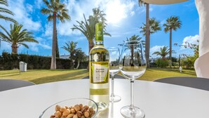 Una botella de vino, dos copas y un bol de frutos secos sobre una mesa con vistas a palmeras y un cielo despejado.