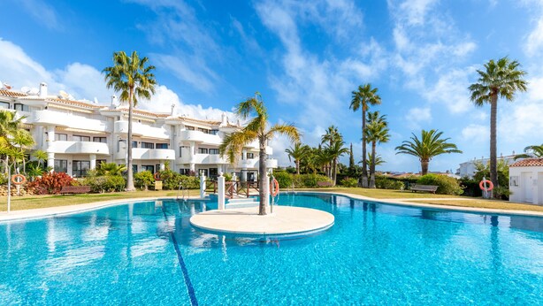 Una piscina azul cristalina con una isla circular en el centro, rodeada de palmeras y un edificio blanco con balcones al fondo.