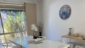 A dining area with a white table and chairs, a clock on the wall, and a view of greenery outside. - House Nelson bay 2min walk to Shops (Nelson Bay)