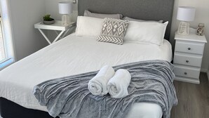A neatly made bed with a grey blanket and two rolled towels, a grey headboard, white nightstands with lamps, and a wall-mounted air conditioner. - House Nelson bay 2min walk to Shops (Nelson Bay)
