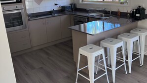 A modern kitchen with a central island, bar stools, built-in appliances, and a window with a view of greenery. - House Nelson bay 2min walk to Shops (Nelson Bay)