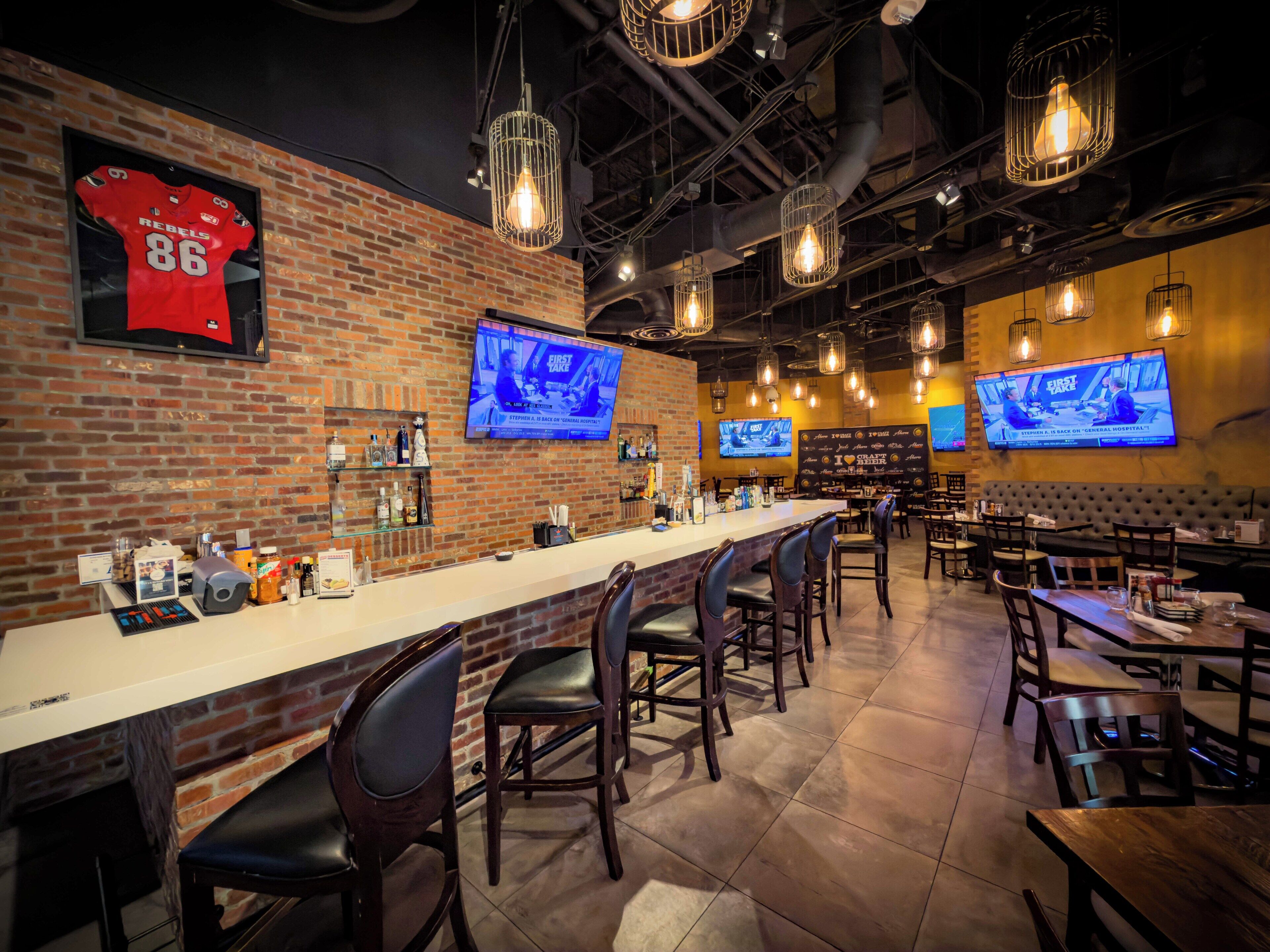 Uma área de bar com paredes de tijolo, várias televisões, luminárias pendentes e banquetas.