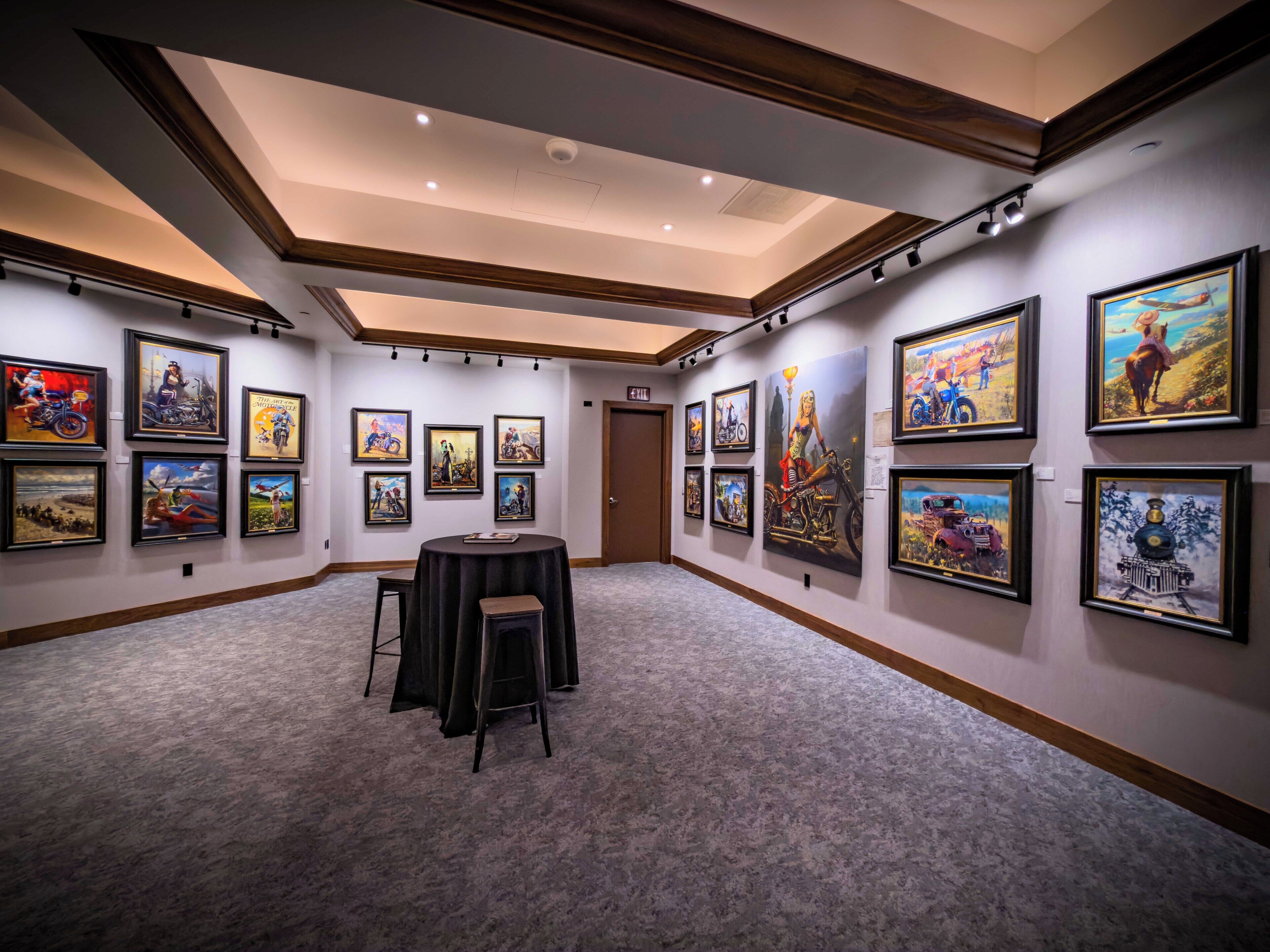 A gallery with framed artworks on the walls, a round table with chairs, and a door in the background.