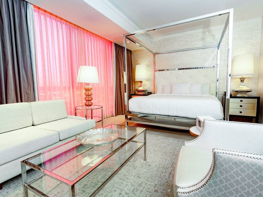 A hotel room with a sofa, a glass-top coffee table, a bed with a canopy, and two bedside tables with lamps.