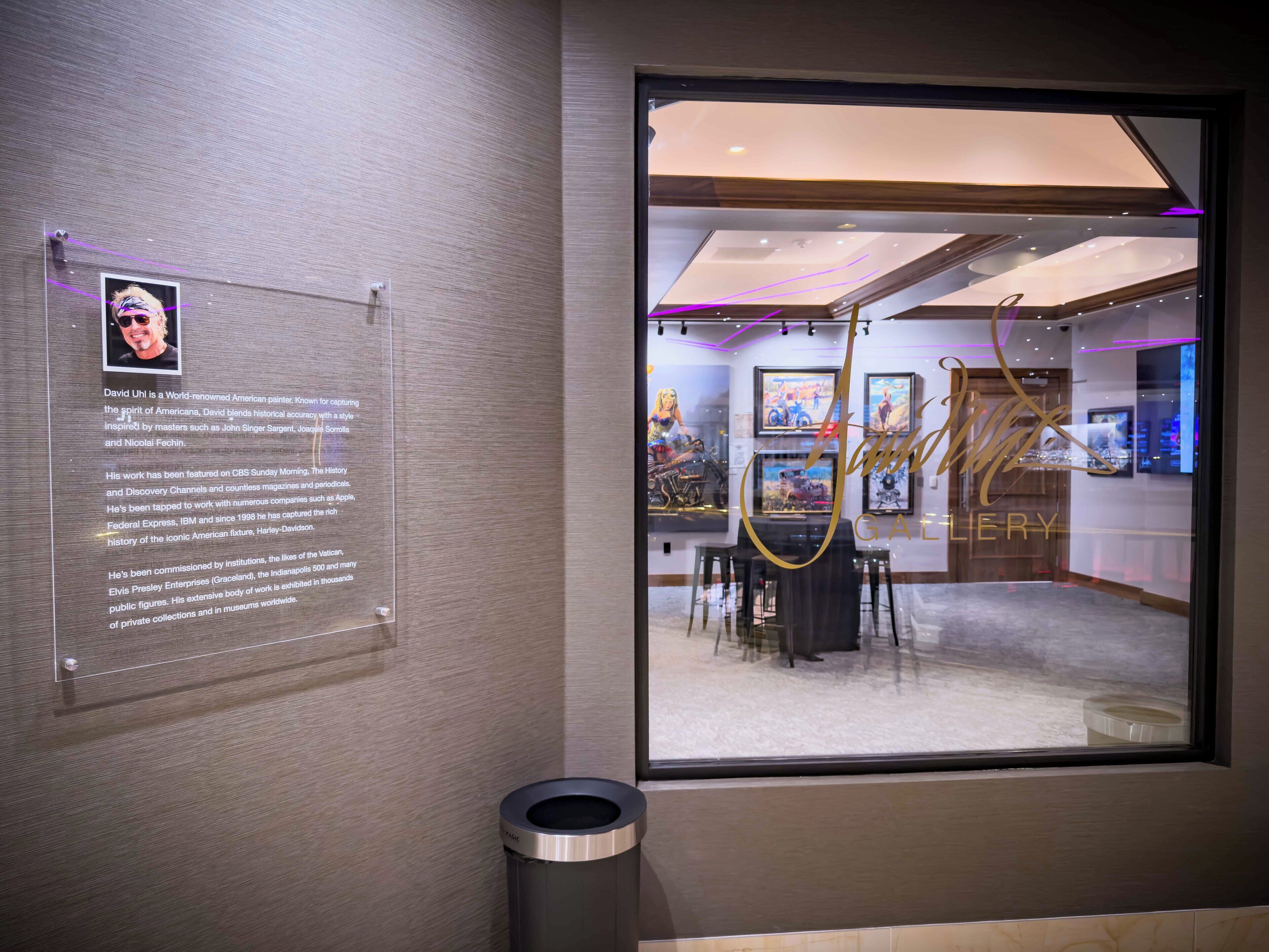 Interior de una galería con una placa informativa enmarcada, un cubo de basura y una vista del interior de la galería a través de una ventana.