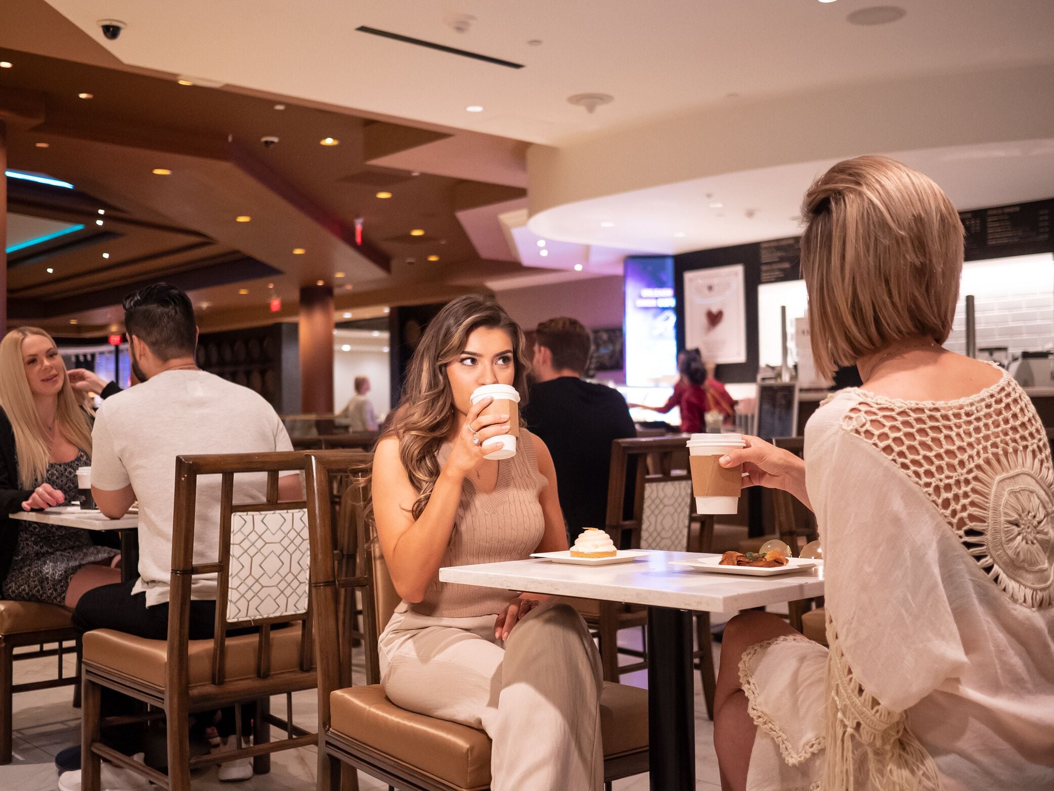 A modern indoor dining area with wooden chairs and tables, a visible ceiling with recessed lighting, and a person holding a coffee cup.