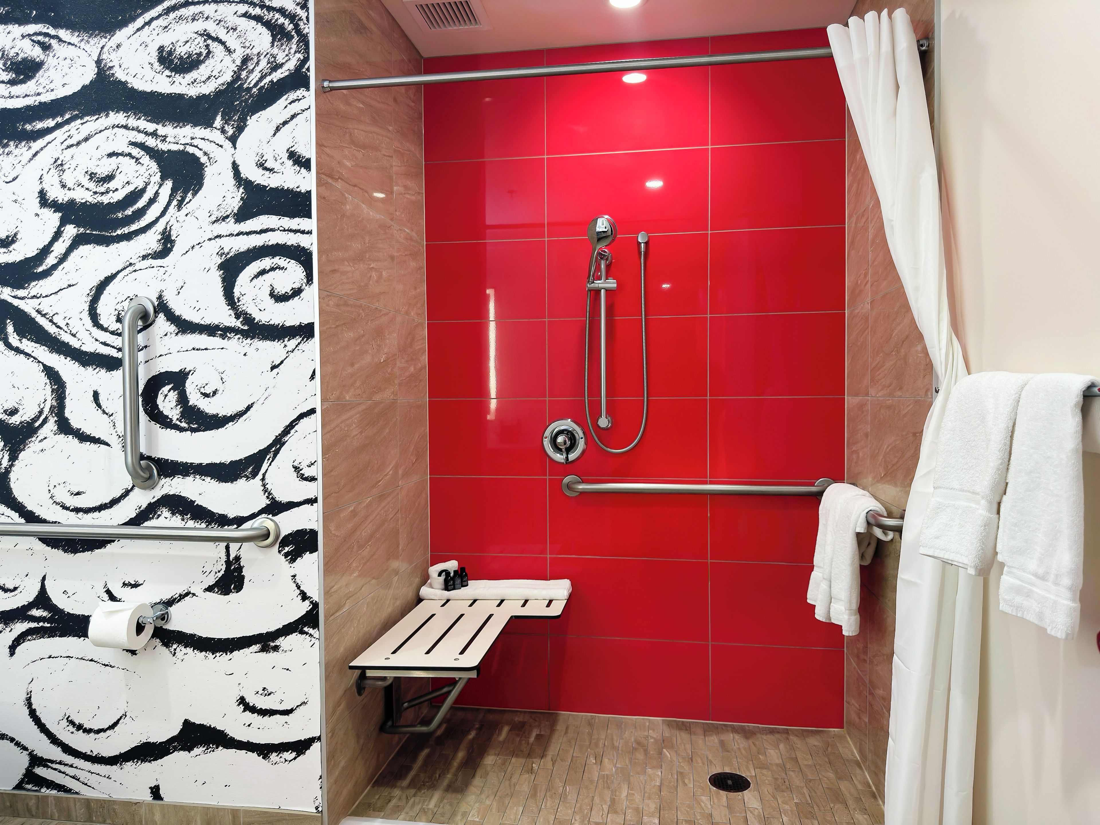 Un baño con pared de azulejos rojos, una ducha con cabezal de ducha manual, un perchero para toallas y un dispensador de papel higiénico.