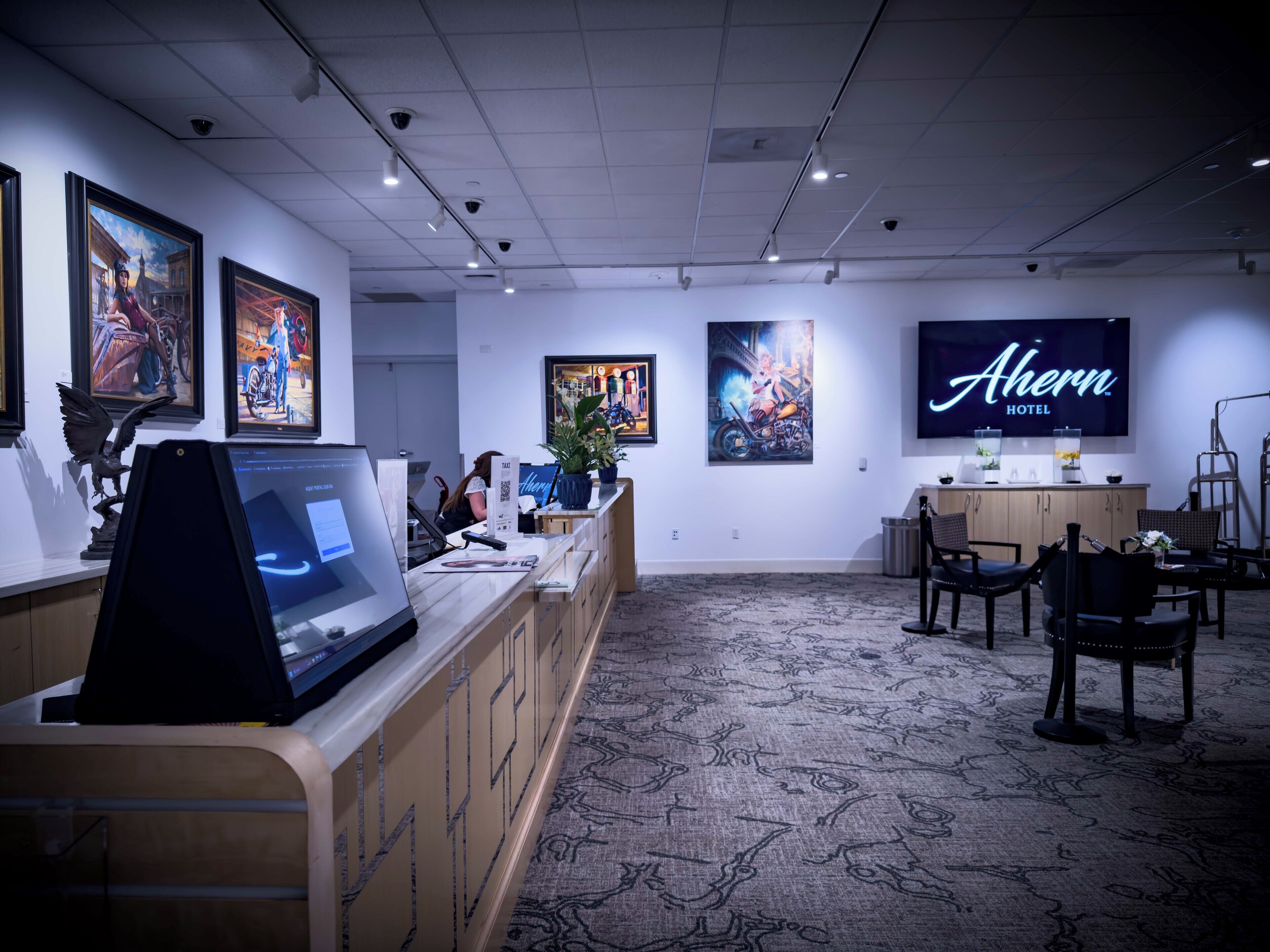 A hotel reception area with artwork on the walls, a large TV displaying the hotel name, and a reception desk with a laptop and a plant.